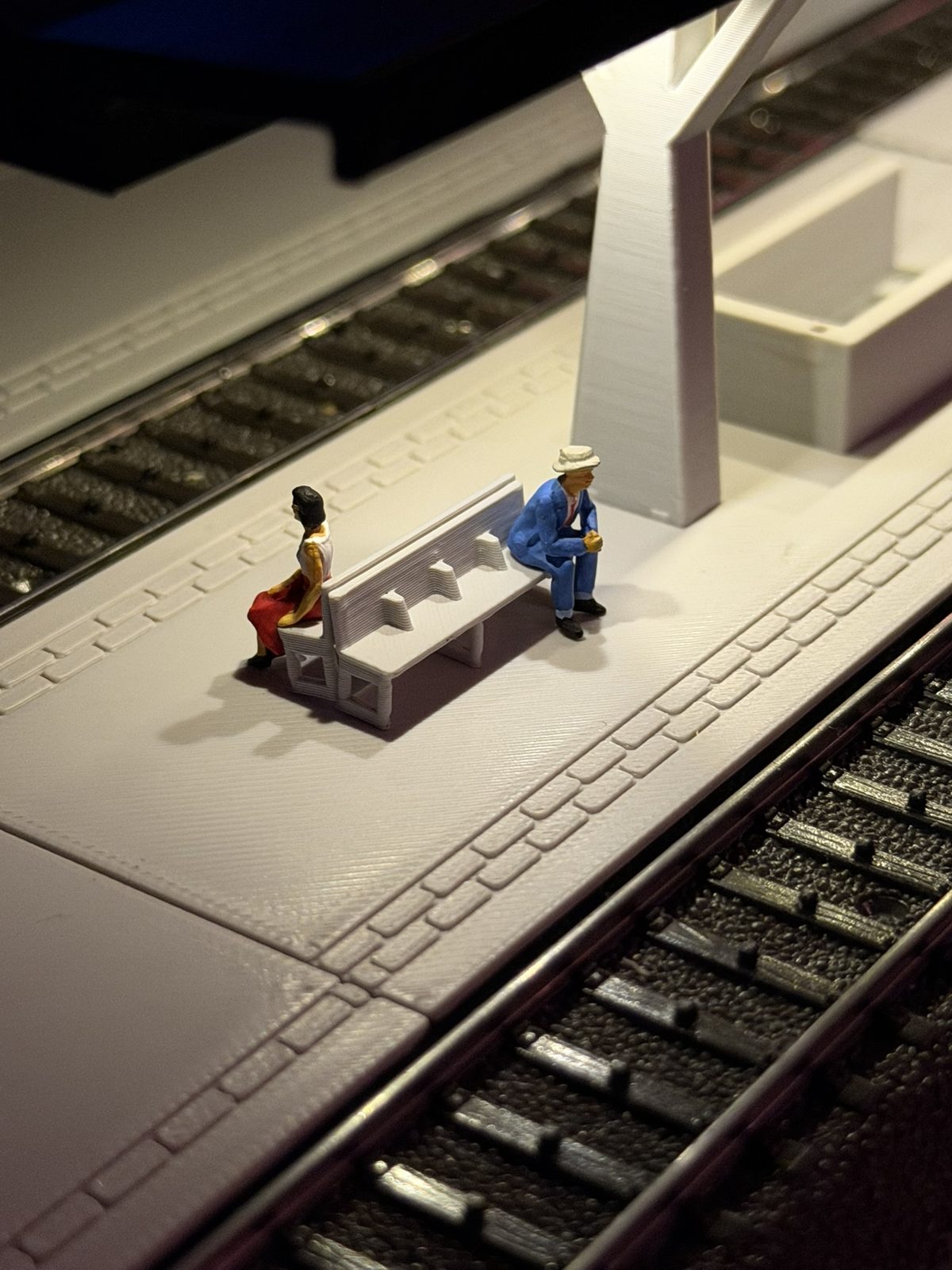 Real print: two figures on a bench under the Y pillar of the V roof station